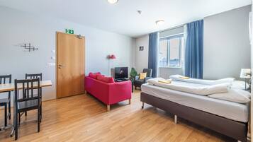 A modern hotel room with a wooden floor, a bed, a red sofa, a TV, and a window with curtains.