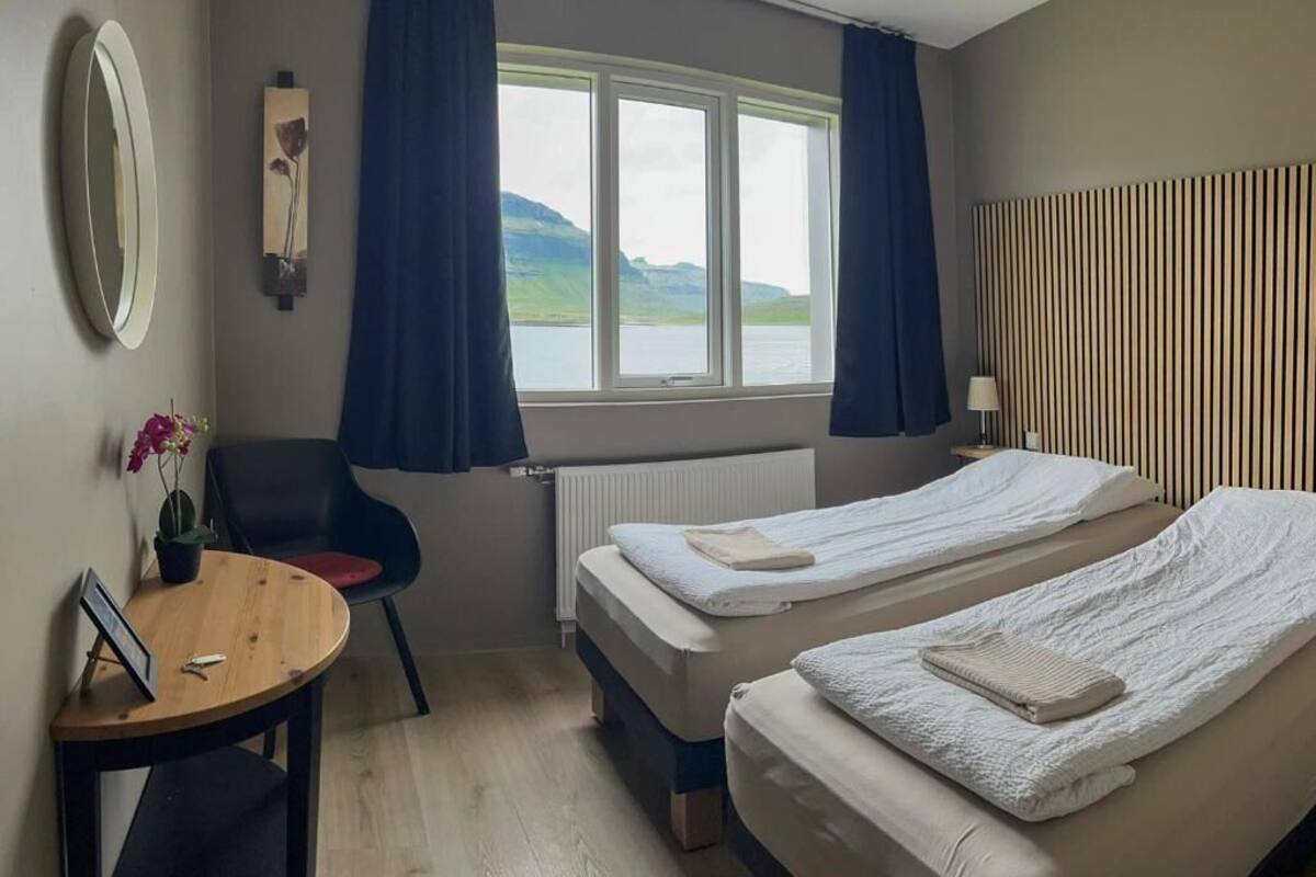 A hotel room with two beds, a desk, a chair, a window with a view of mountains, and a radiator.