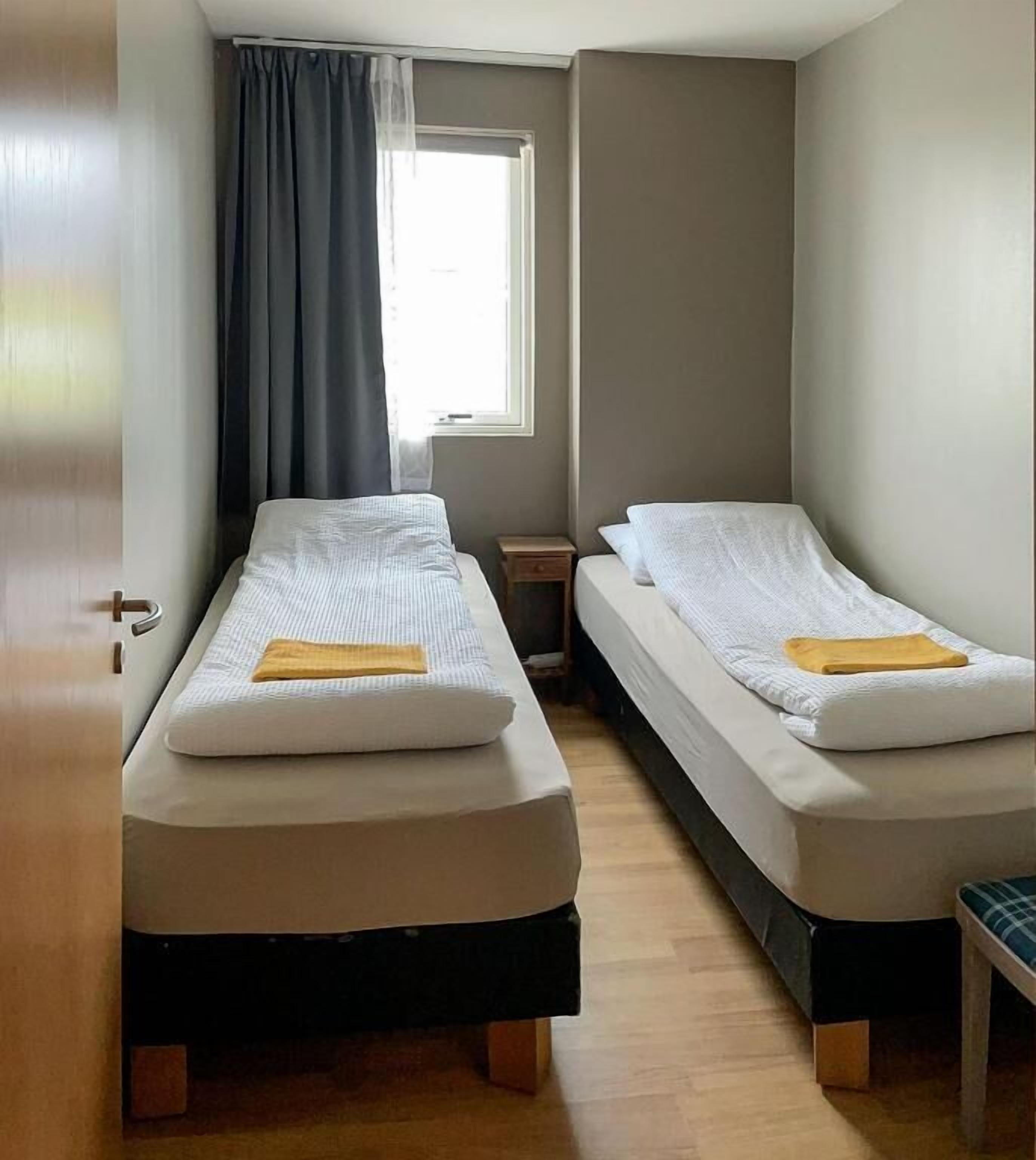 A room with two single beds, a small table, a window with curtains, and a door.