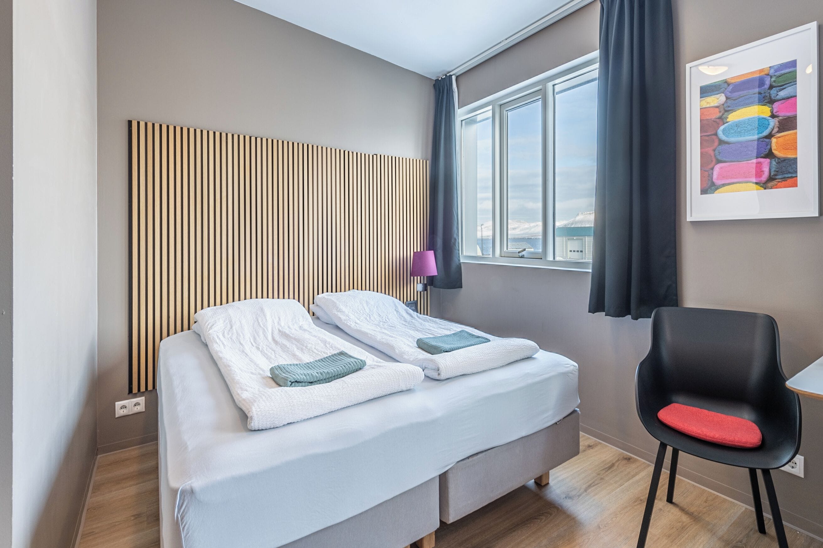 A modern hotel room with a bed, a chair, a window, and a wall-mounted artwork.
