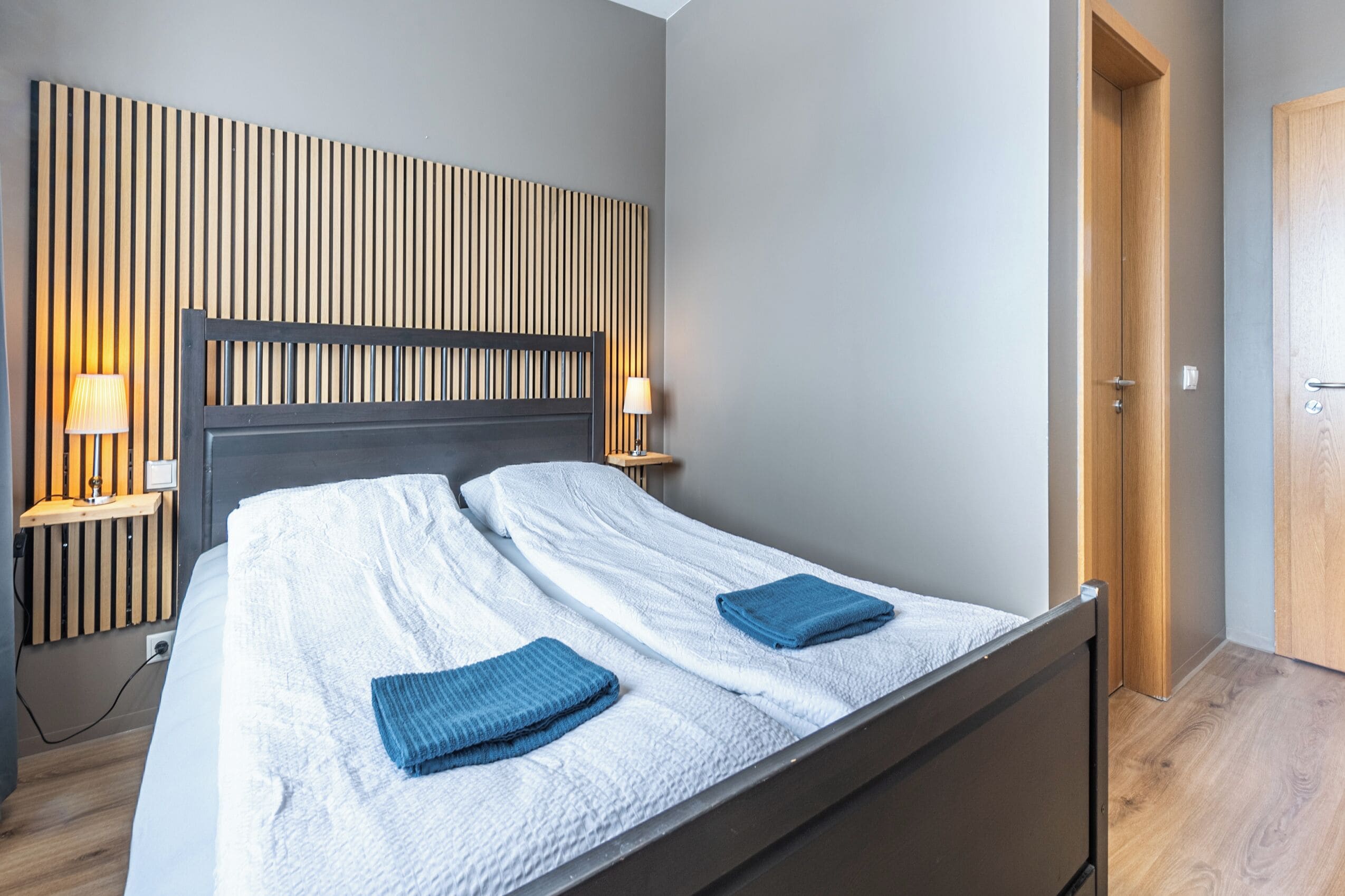A modern bedroom with a wooden headboard, a single bed with a white duvet and two blue towels, bedside lamps, and a wooden door.