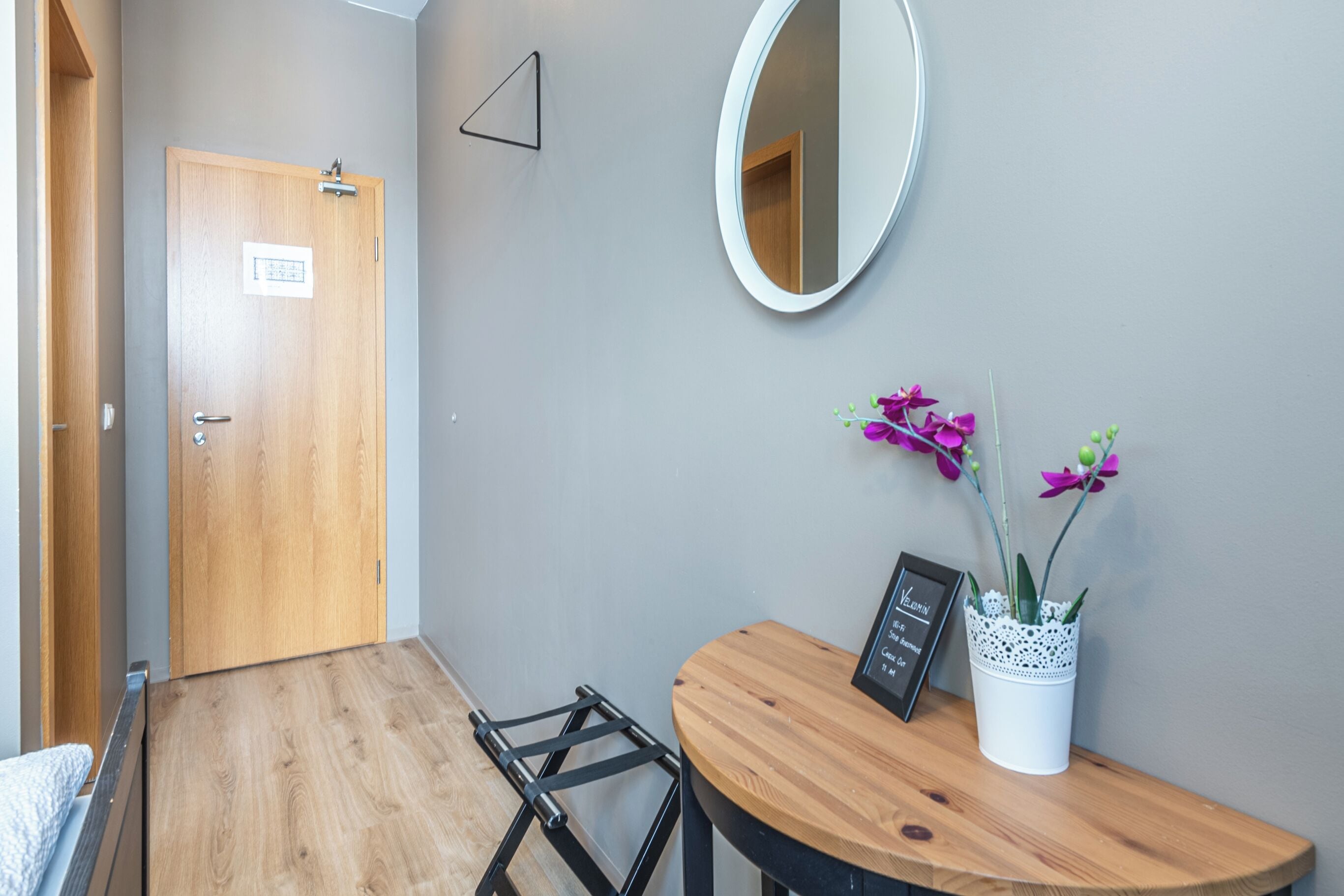 Una habitación con piso de madera, una puerta de madera, una pequeña mesa con un marco de fotos y una planta en maceta, y un espejo en la pared.