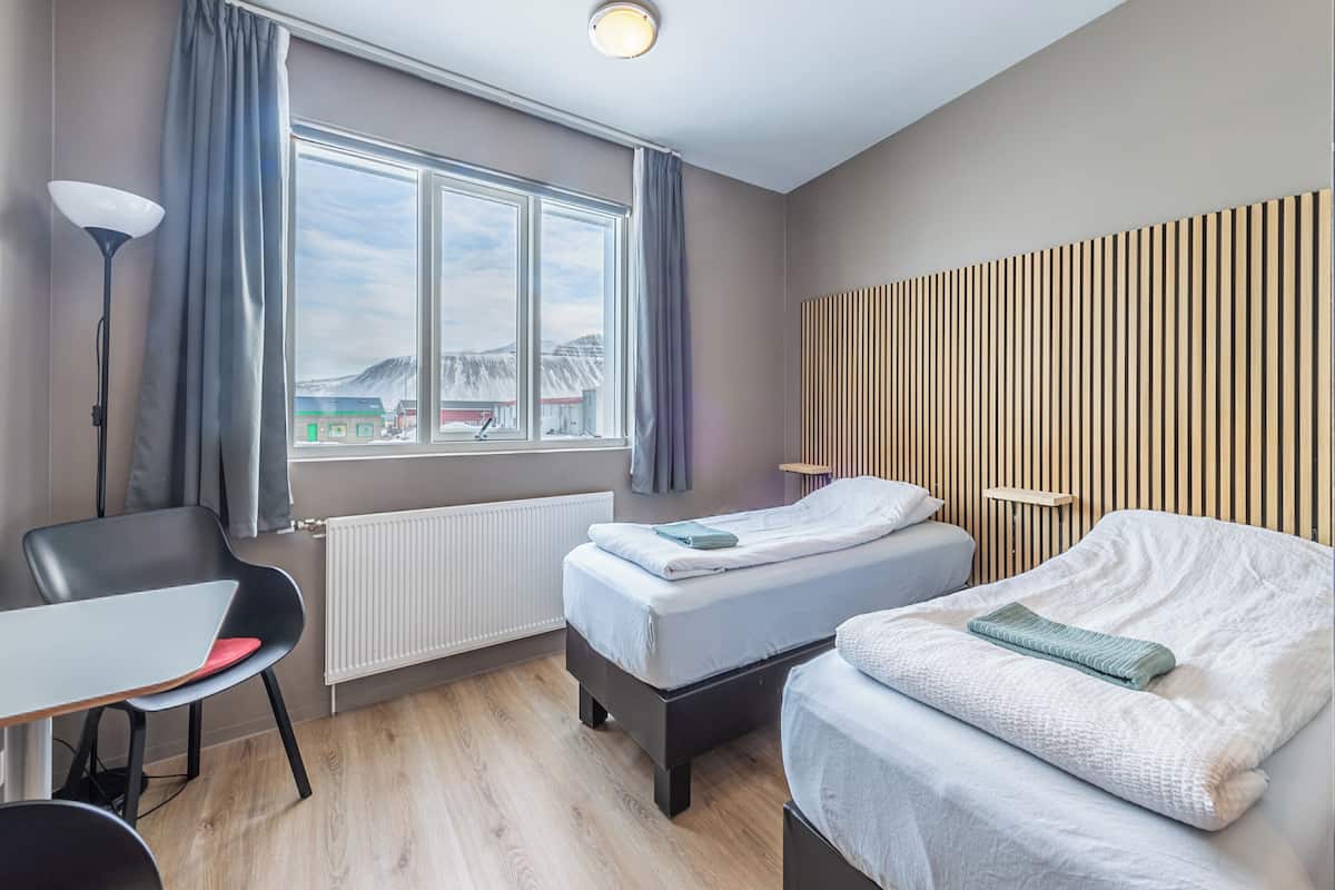 A hotel room with two beds, a desk, a chair, a radiator, and a window with curtains.