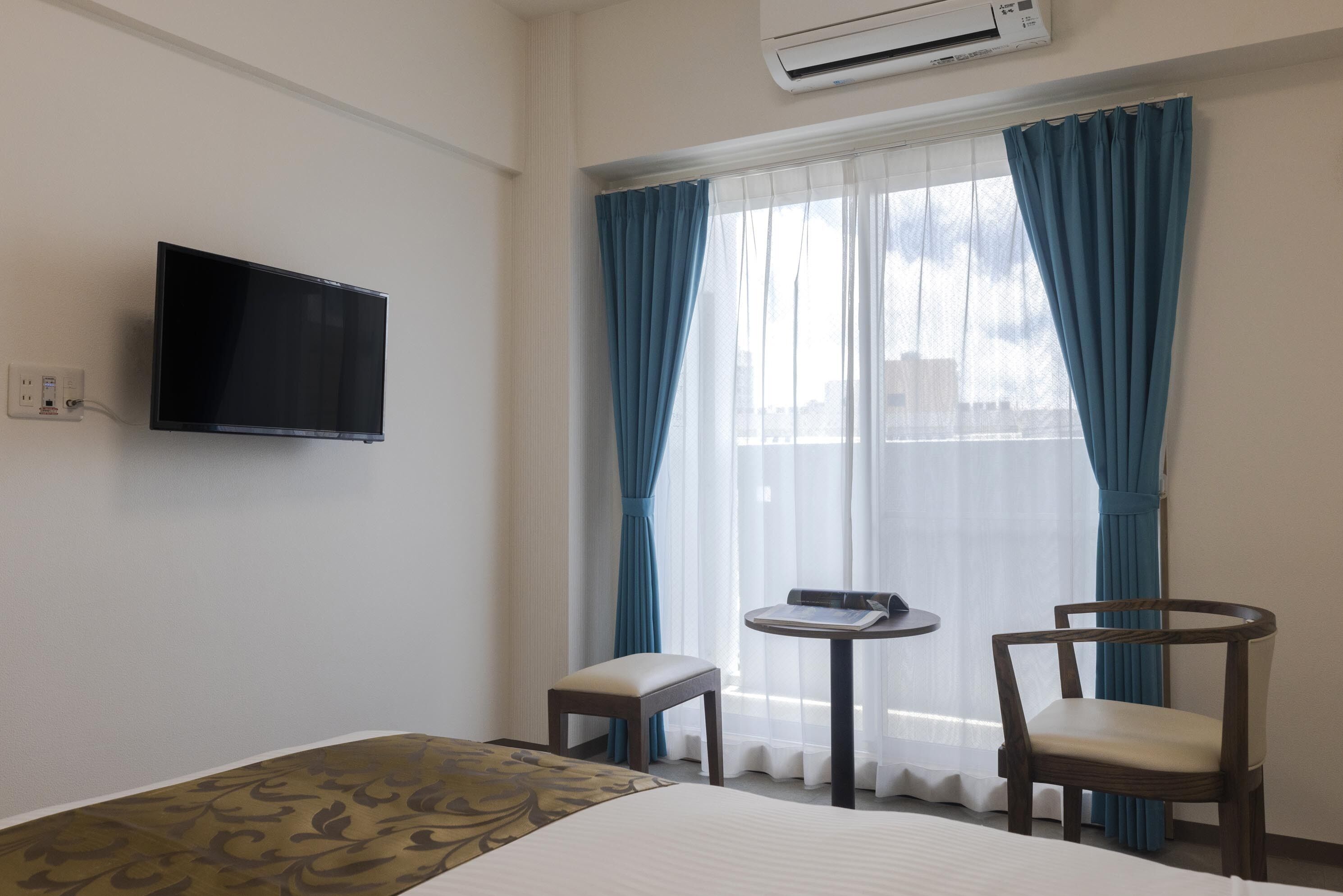 A hotel room with a bed, a TV mounted on the wall, a small table with a chair, and a window with blue curtains.