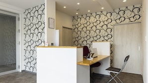 A reception desk with a white counter, a wooden desk, and a folding chair. There is a patterned wall behind the desk and a door to the left. - Mr.KINJO in Miegusuku (Naha)