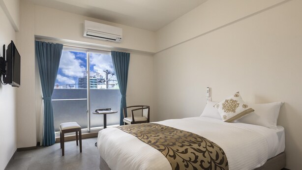 A hotel room with a bed, a chair, a small table, and a view of a balcony with a cityscape. - Mr.KINJO in Miegusuku (Naha)