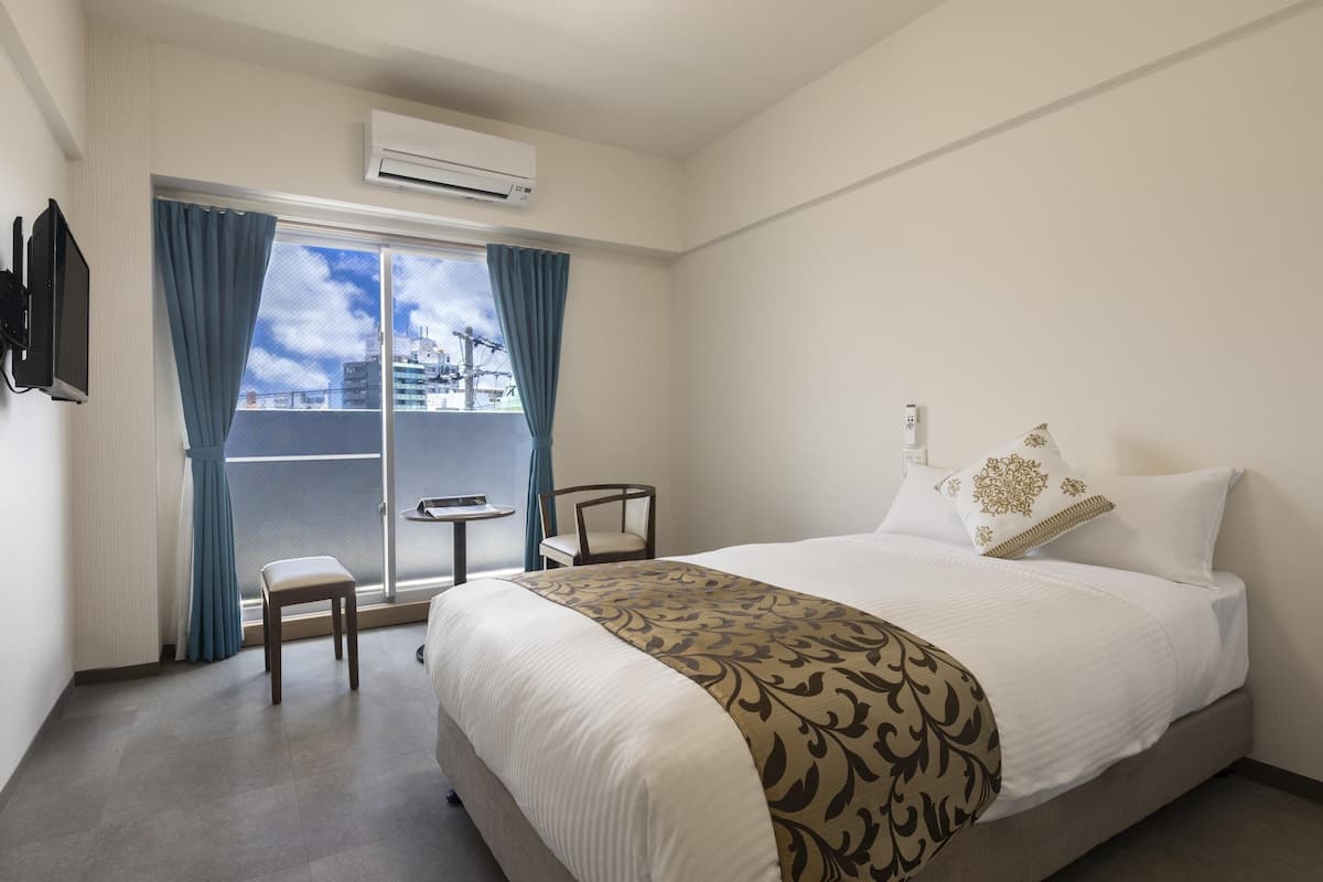 A hotel room with a bed, a chair, a small table, and a view of a balcony with a cityscape.
