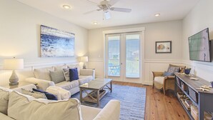 A spacious living room with a ceiling fan, a large window with blinds, a flat-screen TV, and a dining area with wicker furniture. - Grand Isle 302 - Community Pool, Private Beach Access, 30A, Near Seaside! (Seagrove Beach)