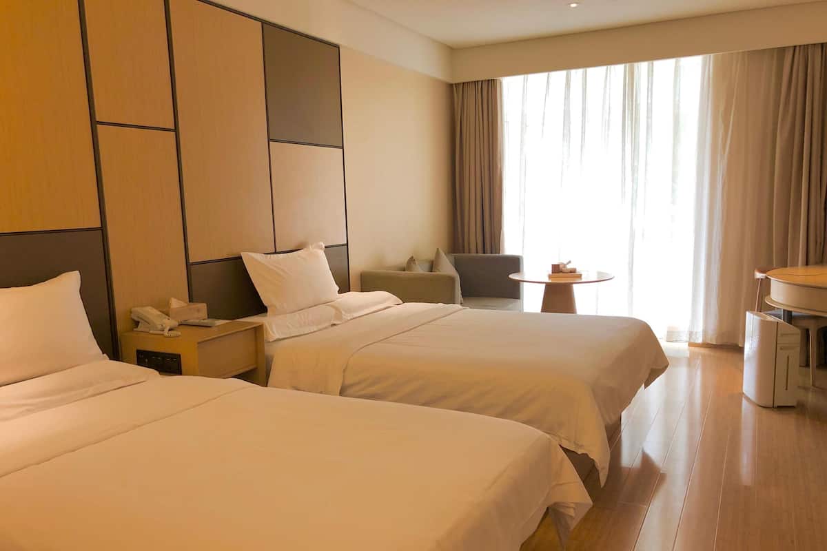 Une chambre d’hôtel avec deux lits, un bureau, une chaise, une petite table et une grande fenêtre avec des rideaux.
