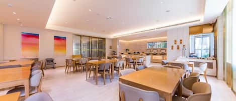 Un comedor moderno con varias mesas y sillas, una cocina al fondo y ventanas grandes.