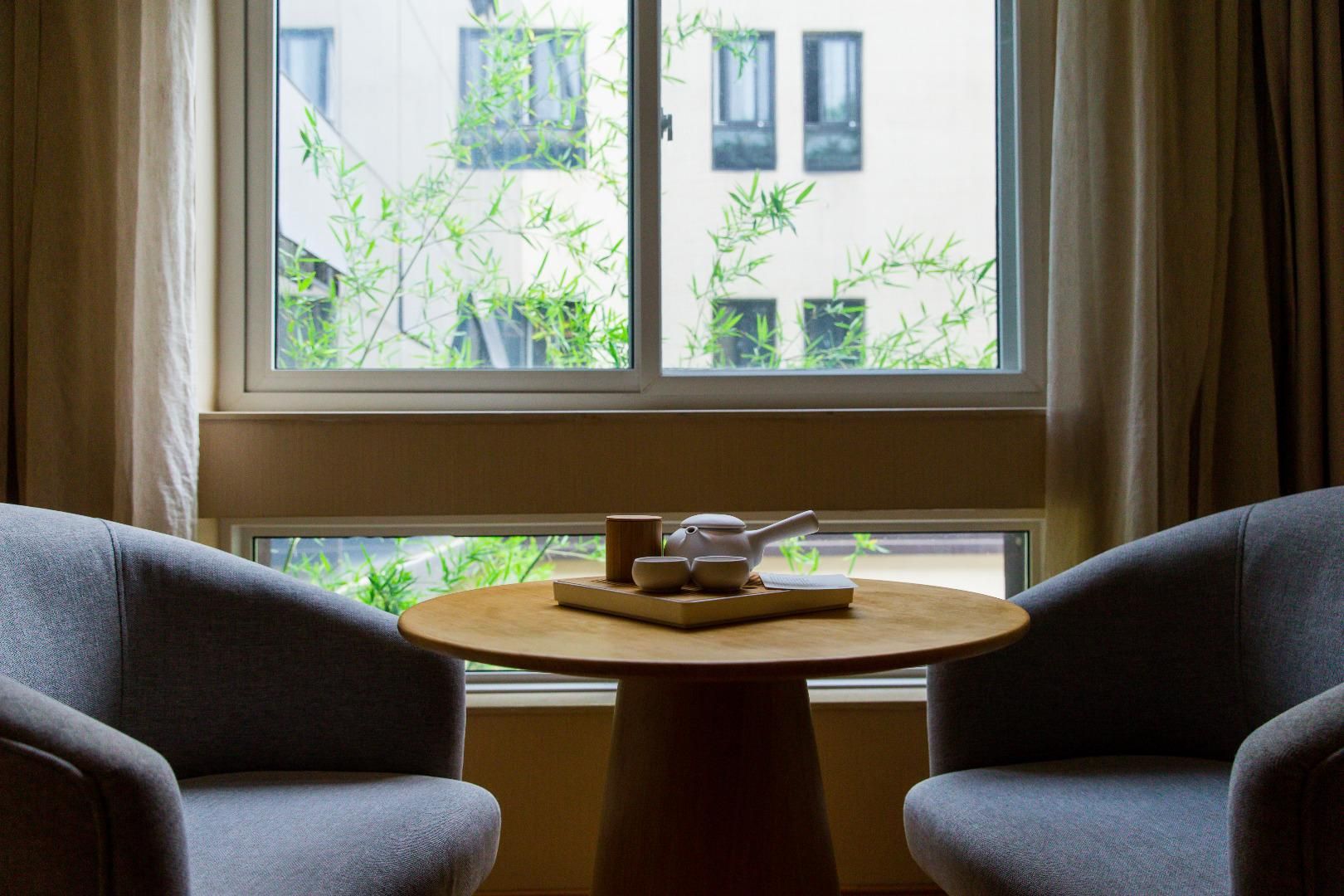 A room with a round wooden table, two grey armchairs, and a window with bamboo plants outside.