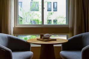 Una habitación con una mesa redonda de madera, dos sillones grises y una ventana con plantas de bambú afuera.