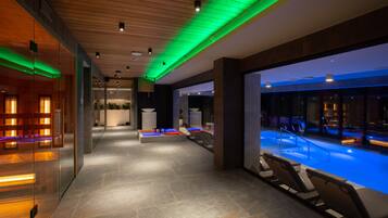 A modern indoor swimming pool area with lounge chairs, a reception desk, and a glass wall.