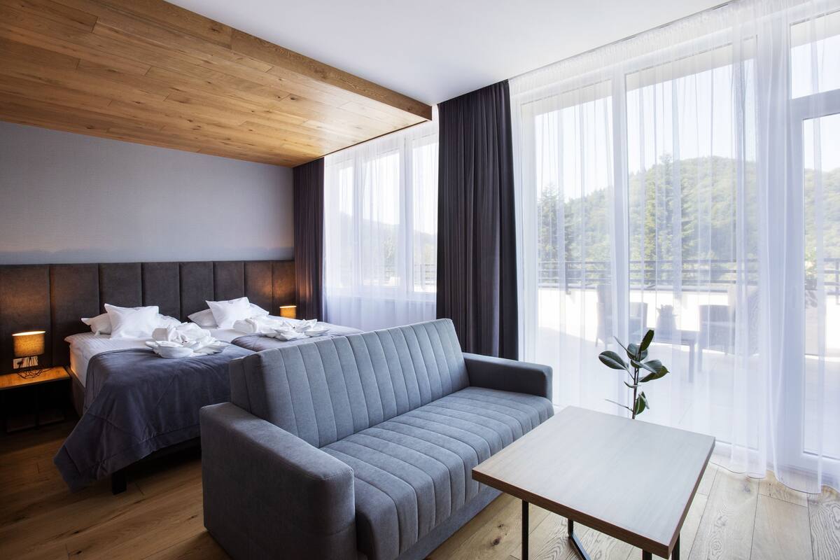 A modern hotel room with a wooden ceiling, a bed with a headboard, a grey sofa, a wooden table, and large windows with sheer curtains.