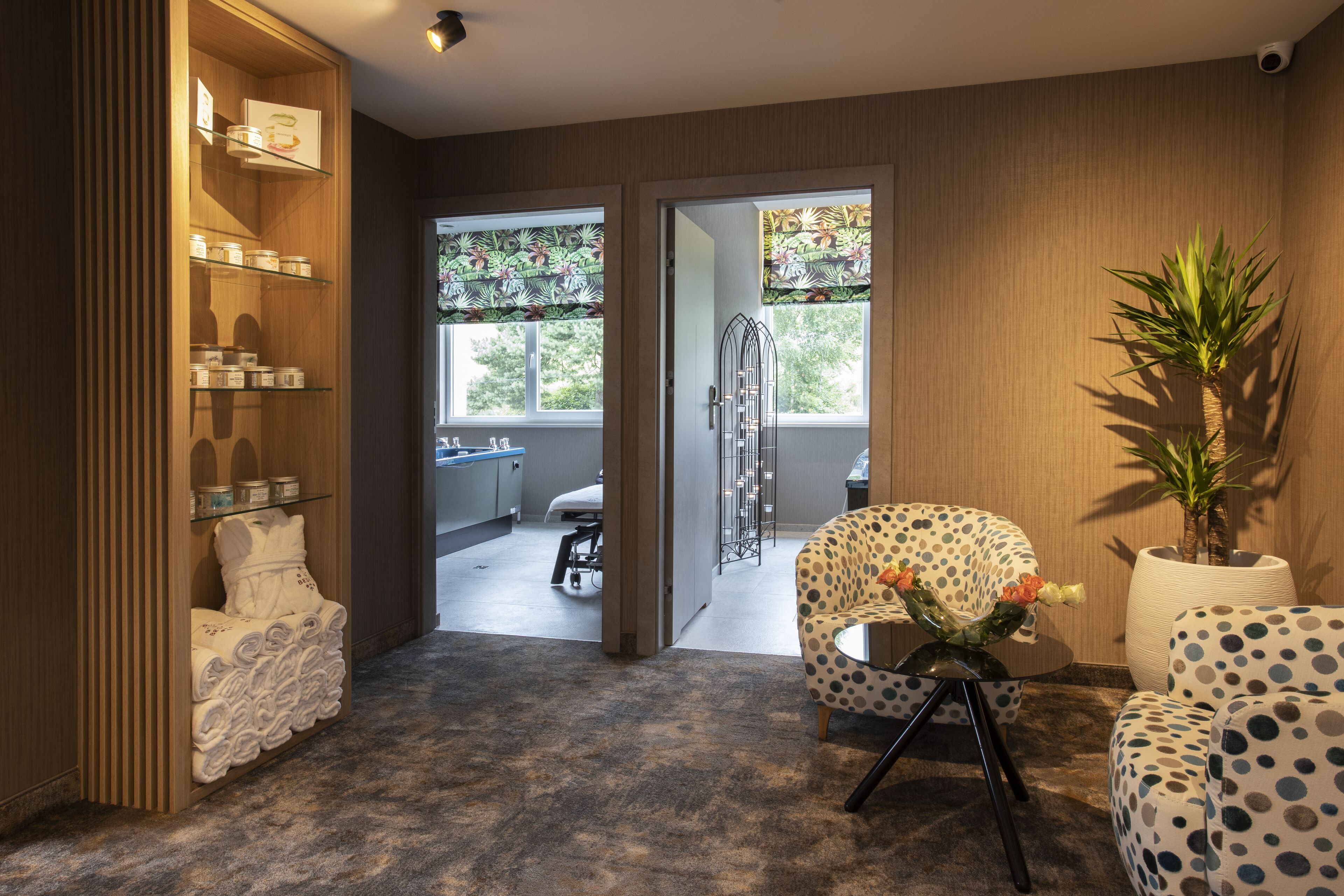 Una habitación de hotel moderna con un armario de madera, un sillón a lunares, una planta en maceta y vistas al baño a través de una puerta de vidrio.