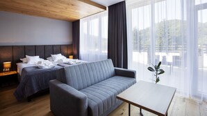 A modern hotel room with a wooden ceiling, two beds with grey bedding, a grey sofa, a wooden table, and large windows with sheer curtains. - Hotel Beskid Balneo Medical Resort & SPA (Piwniczna-Zdroj)