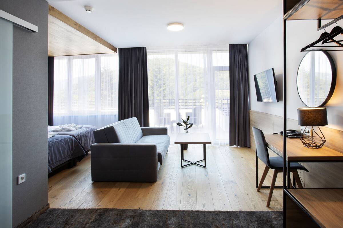 A modern hotel room with a sofa, a dining table, a TV mounted on the wall, and large windows.