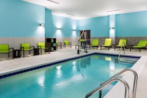 Un área de piscina con piso de baldosas azules, una piscina con escalera metálica y sillas verdes a lo largo del borde.