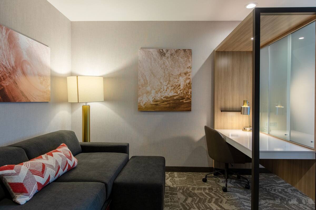 A modern hotel room with a grey sofa, a desk, and a glass partition.