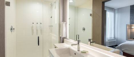 A modern bathroom with a glass-enclosed shower, a large white sink, and a mirror reflecting a bedroom with a bed and window.