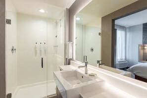 Un baño moderno con ducha envuelta en vidrio, un lavamanos grande de color blanco y un espejo que refleja un dormitorio con cama y ventana.