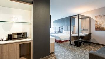 A hotel room with a kitchenette, a bed with white linens, a desk with a chair, and a large abstract painting on the wall.