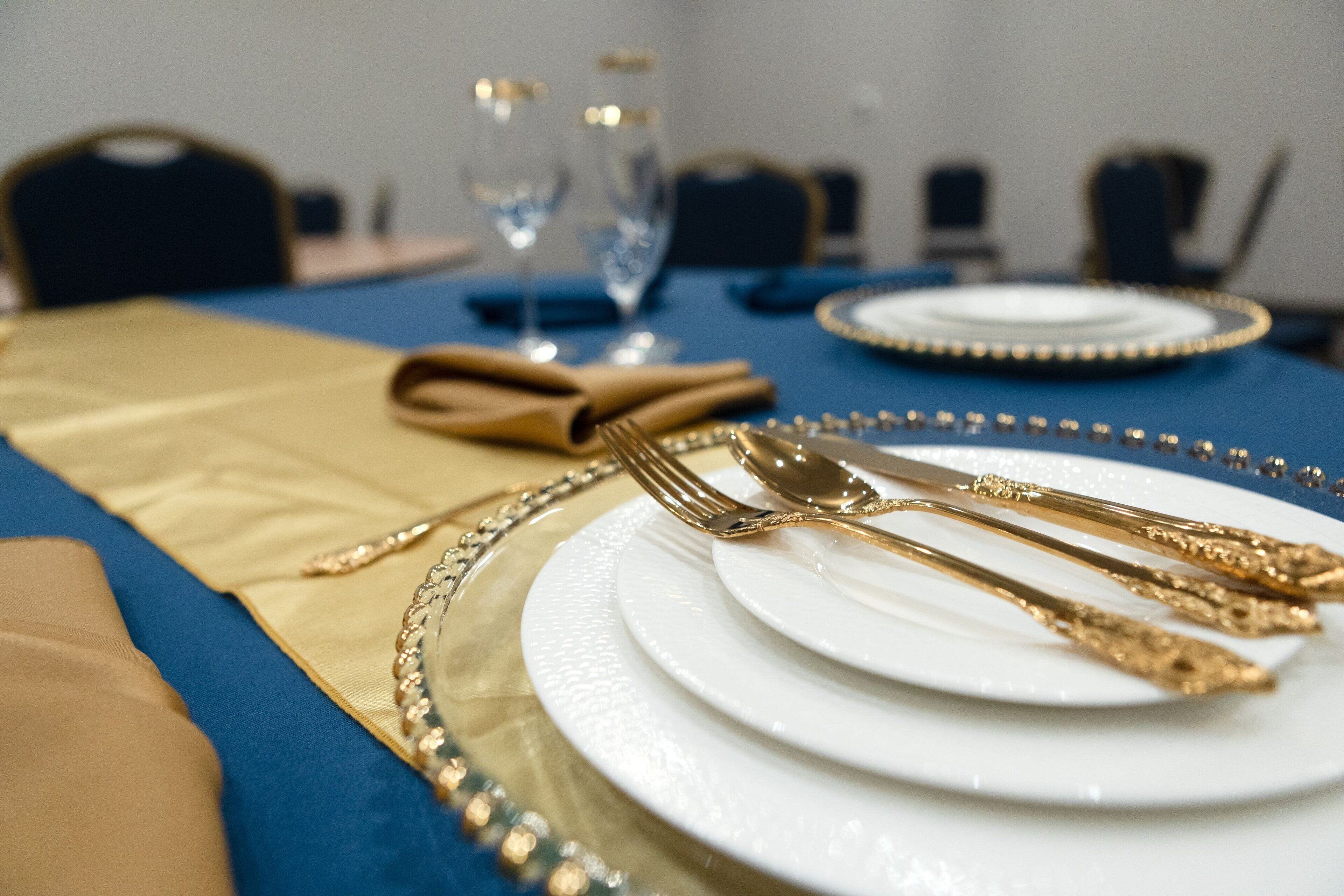 Une table dressée avec des couverts en or et des assiettes blanches, une nappe bleue et une serviette en tissu dorée.