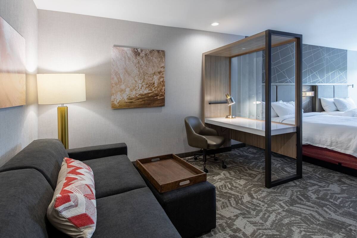 A modern hotel room with a sofa, a desk, a bed, and a large abstract painting on the wall.