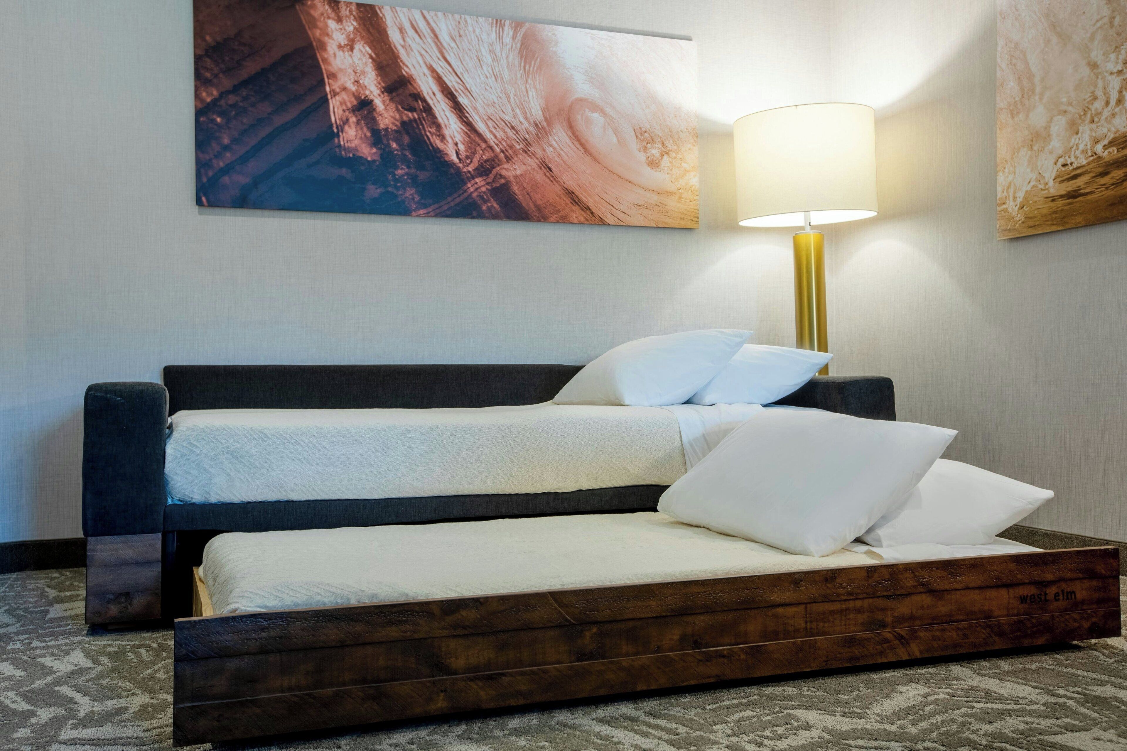Una cama doble con estructura de madera, cabecera oscura y ropa de cama blanca. Se observa una lámpara de pared con base dorada y pantalla blanca.