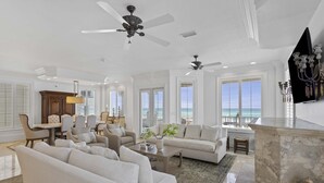 A spacious living room with a large white sofa, a coffee table, and a fireplace. There are multiple ceiling fans and large windows offering a view of the ocean. - Kings View|Private Beach Access|Gulf Front Home with Stunning Gulf Views (Destin)