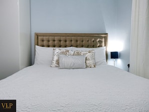 Relax in a spacious room with a large bed and comfortable pillows, featuring a tufted headboard and a bedside table with a lamp.