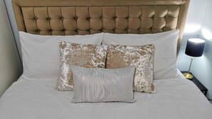 Una cama con cabecera capitonada, dos almohadas estampadas y una lámpara de noche.