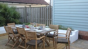 Un espacio de comedor al aire libre con una mesa y sillas de madera, rodeado por una terraza de madera y un bajo muro de ladrillo.