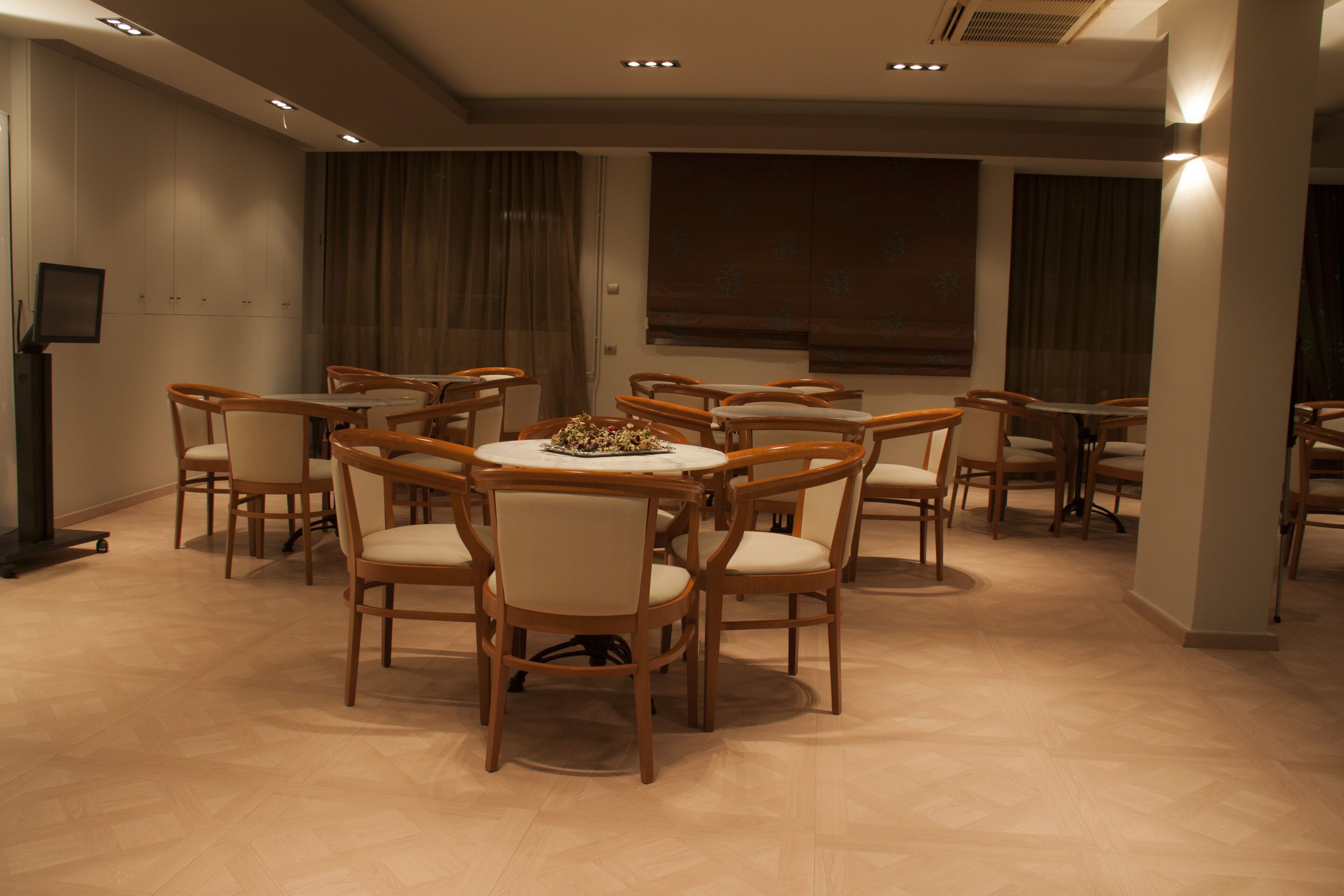 Un salón de conferencias con varias mesas y sillas, un televisor de pantalla plana y grandes ventanas con cortinas.