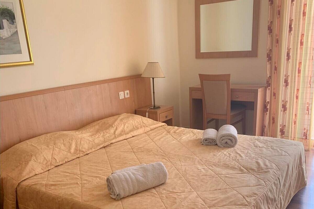 Una cama bien hecha con cubrecama acolchada, una lámpara de noche y una silla en una habitación con suelo de madera y un espejo en la pared.