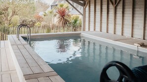 An indoor swimming pool with wooden beams, a metal ladder, and a view of a garden with various plants. - Stone farmhouse for 6 people with heated swimming pool (Trélévern)