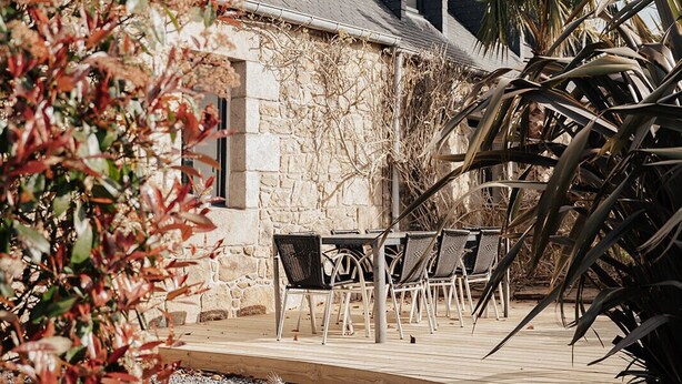 A stone building with a sloped roof, outdoor seating, and a palm tree. - Stone farmhouse for 6 people with heated swimming pool (Trélévern)