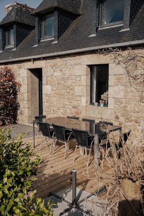 Un bâtiment en pierre doté d’une terrasse en bois, d’un espace extérieur aménagé pour s’asseoir et d’une fenêtre ornée d’une plante.