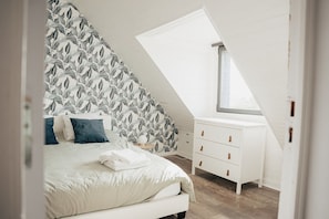 Une chambre avec un plafond en pente, un lit avec une literie blanche, une commode et une fenêtre.
