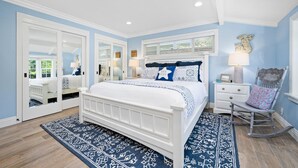 Un dormitorio con una cama blanca, ropa de cama con estampado azul y blanco, una alfombra azul y blanca, un sillón mecedora y un aparador con espejo.