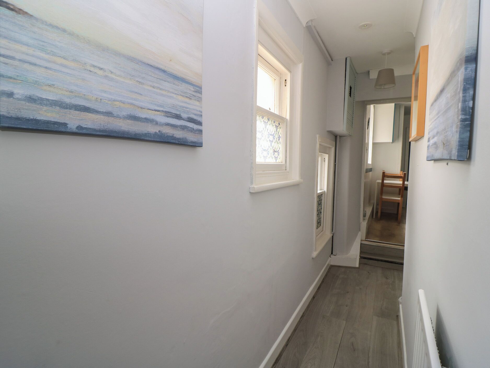 A hallway with a wooden floor, white walls, and a painting on the left.