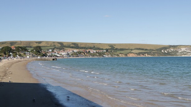A coastal beach with a sandy shore, a clear blue sky, and a town in the background. - Still Waters (Swanage)