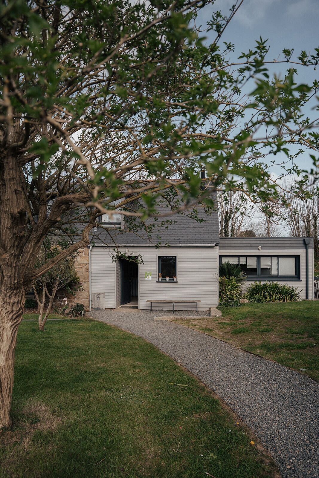 Una casa con camino de grava, un banco de madera y un árbol en primer plano.