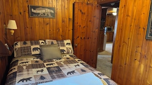 Um quarto com painéis de madeira, uma cama com padrão de urso acolchoado, uma placa com a inscrição 'Hibernation Station' e uma porta que dá para uma área de cozinha.