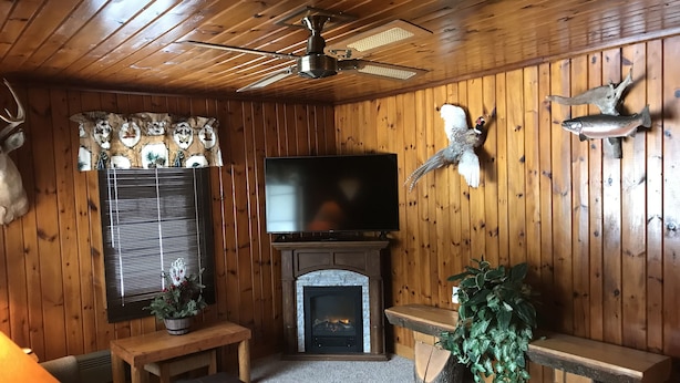 Interior acolhedor de uma cabana com lareira, paredes de madeira, ventilador de teto, televisão e chifres de animais pendurados.