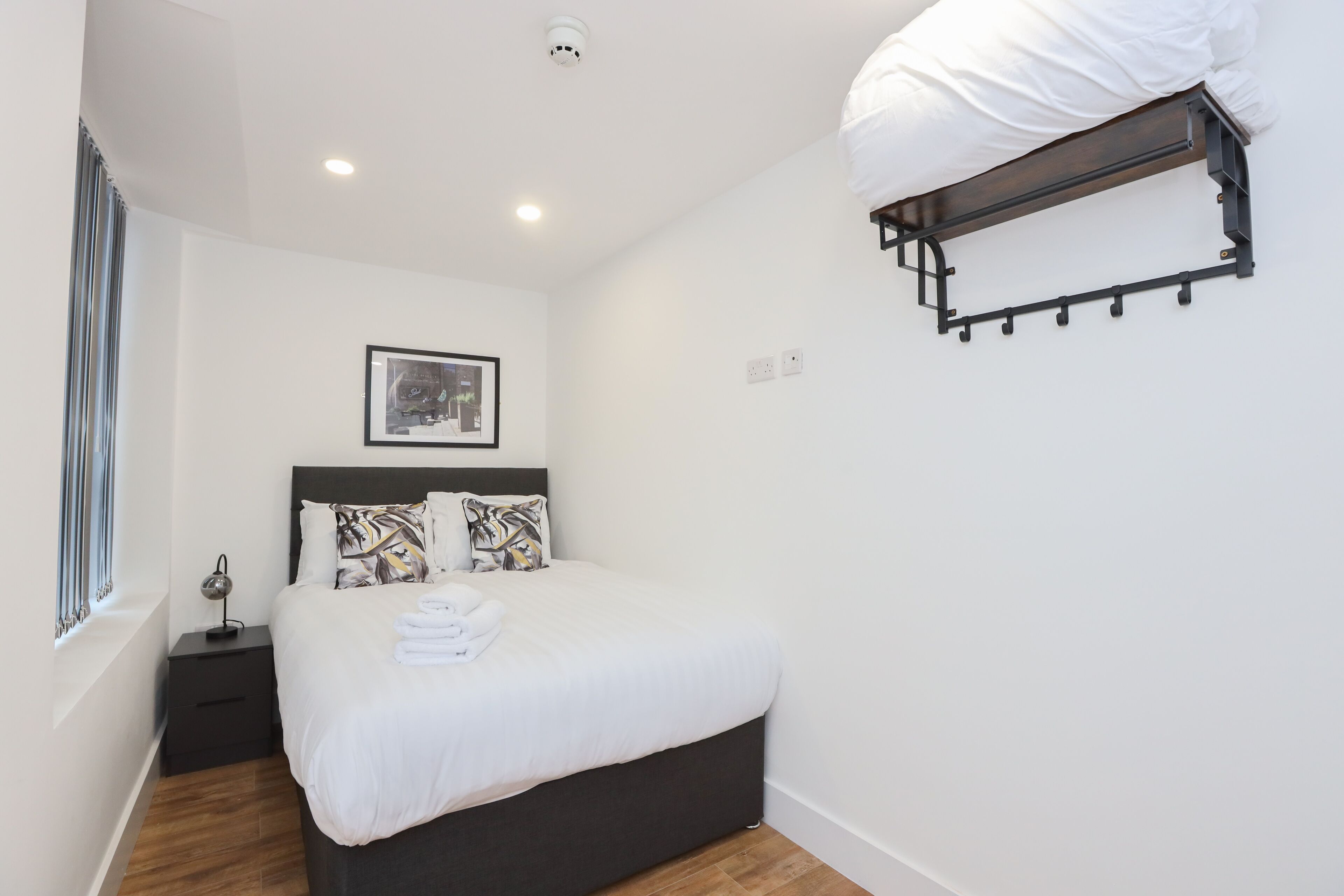 Un lit bien fait, avec une literie blanche, une tête de lit agrémentée d’un coussin décoratif, une table de chevet avec une lampe et un porte-manteau fixé au mur.