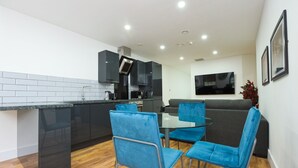 A modern kitchen with black cabinets, a built-in oven, and a dining area with blue chairs and a glass table. - Wood Street Apartments (Liverpool)