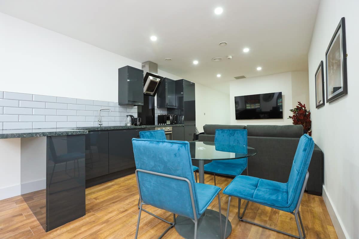 Una cocina moderna con armarios negros, horno integrado y un comedor con sillas azules y una mesa de vidrio.