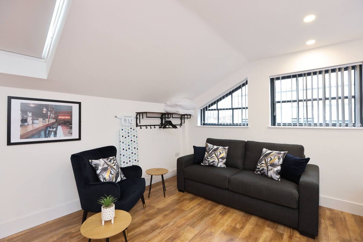 Una sala de estar moderna con un sofá gris oscuro, una mesita redonda pequeña y un sillón negro. Se observa un cuadro en la pared, una pequeña planta en maceta y una ventana con barrotes.