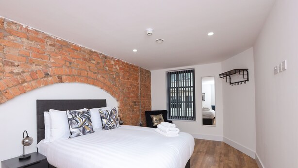 A modern bedroom with a brick accent wall, a large bed with white linens, a nightstand, a mirror, and a window with blinds. - Wood Street Apartments (Liverpool)